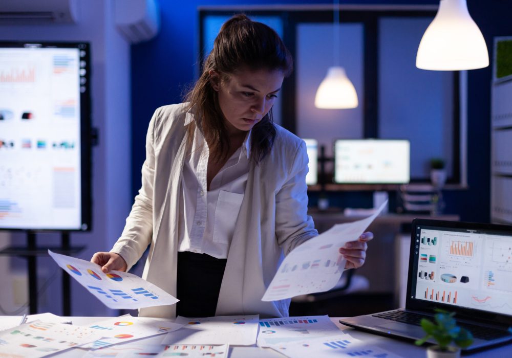 Busy business woman looking on financial reports, checking statistics graphs standing at desk late at night in start-up office. Manager using social network working on marketing project